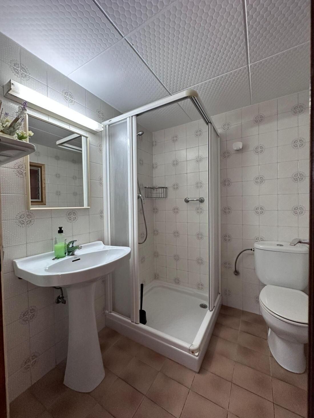 Baño con lavabo de pedestal blanco, ducha con mampara de cristal e inodoro. Azulejos blancos y beige en paredes y suelo.