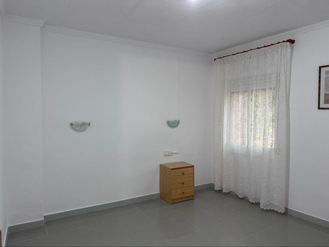 Habitación blanca vacía con una pequeña mesita de noche de madera y una ventana con cortinas transparentes.