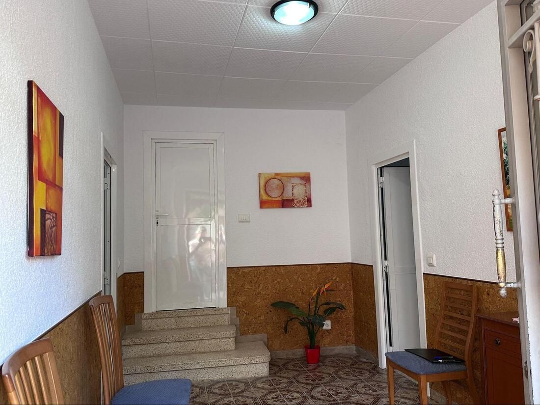 Pasillo con escaleras, puertas, obras de arte y sillas. Paredes blancas, detalles de azulejos marrones y suelo estampado.