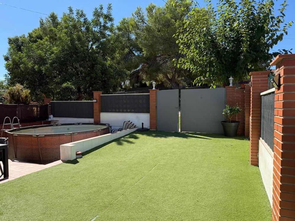 Patio trasero con césped artificial, piscina, portón gris y pilares de ladrillo.