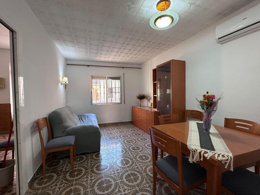 Salón pequeño con sofá, mesa de comedor y armario. Tonos marrones y blancos.