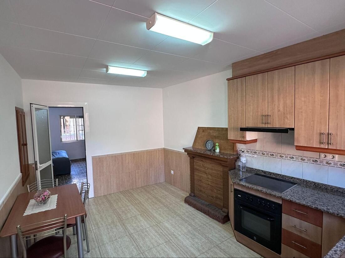 Cocina con armarios de madera, horno y mesa, que da a un pasillo con un dormitorio.