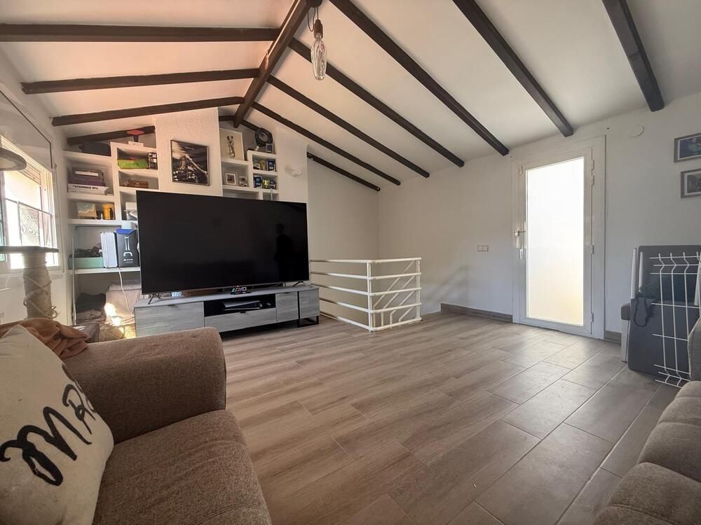 Sala de estar con TV, estanterías, barandilla blanca y suelo de madera. Sofá marrón con cojines.