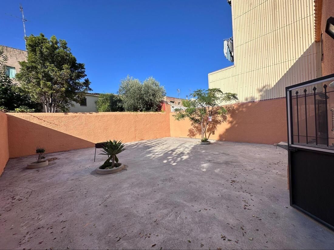 Un pequeño patio trasero, casi desierto, con una pared color coral bajo un cielo azul brillante. Un árbol crece a la derecha.