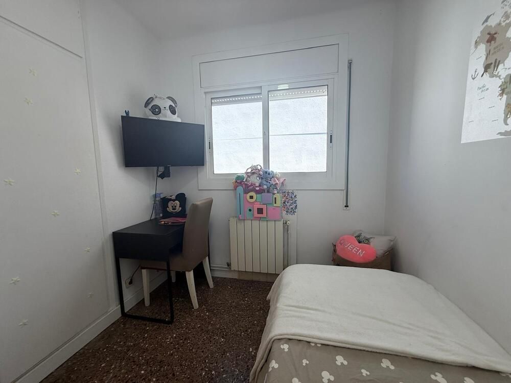 Dormitorio pequeño con cama, escritorio, televisor y ventana. Paredes blancas, suelo oscuro y decoración variada.
