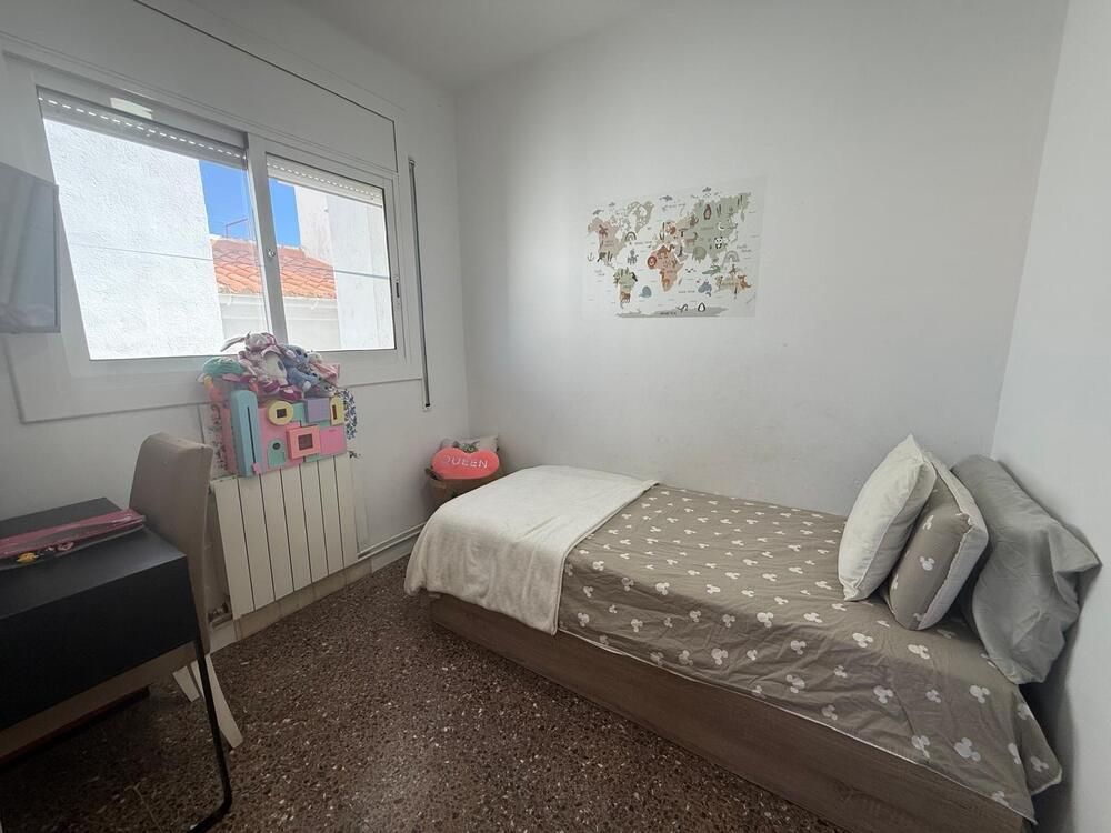 Un dormitorio pequeño con una cama individual, un escritorio y una ventana. Las paredes son blancas y el suelo moteado.