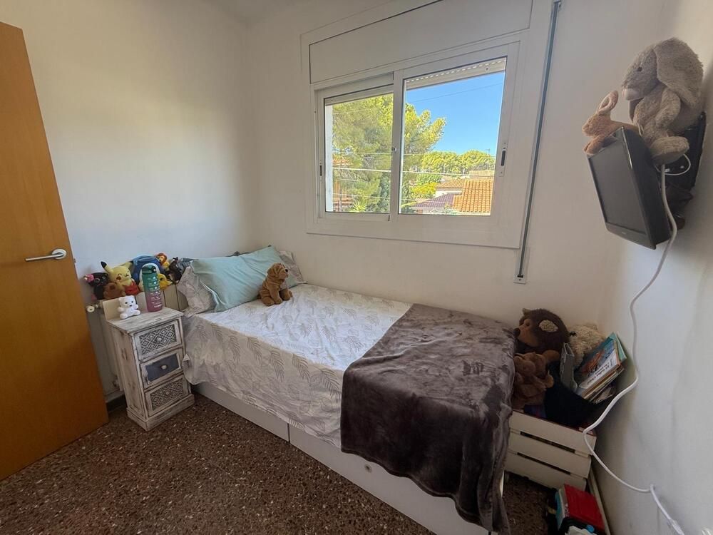 Dormitorio pequeño con cama individual, ventana, mesita de noche y juguetes. Tonos marrones y blancos con luz natural.