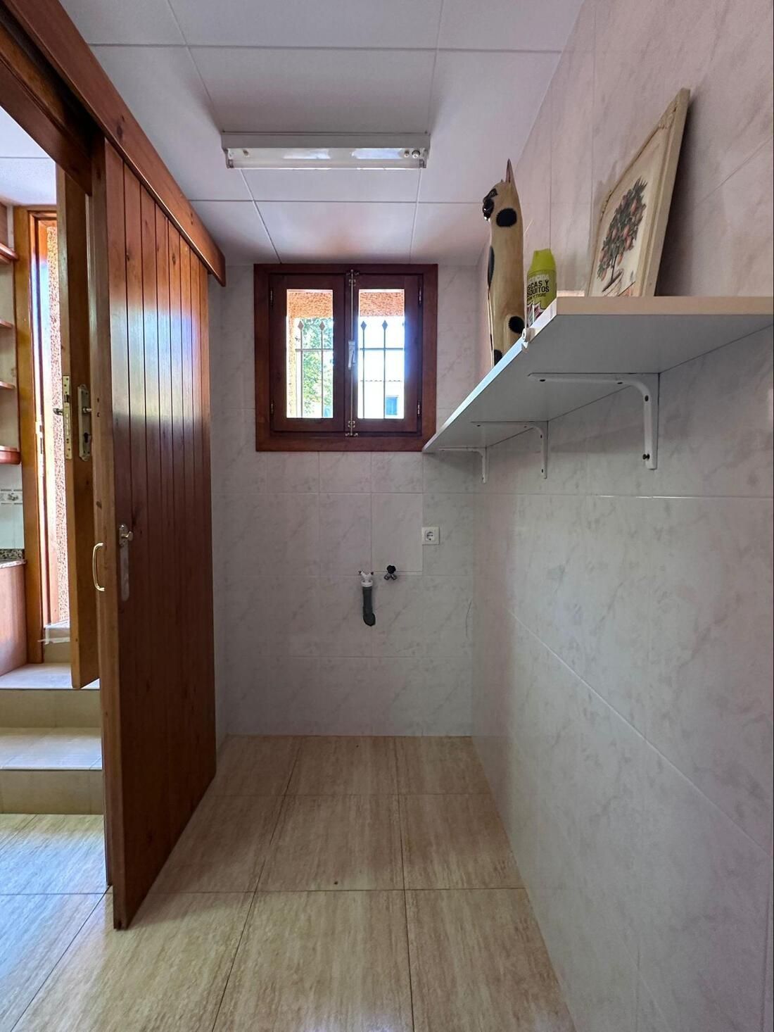Un pequeño cuarto de servicio con azulejos, ventana, estante y puerta corredera de madera.