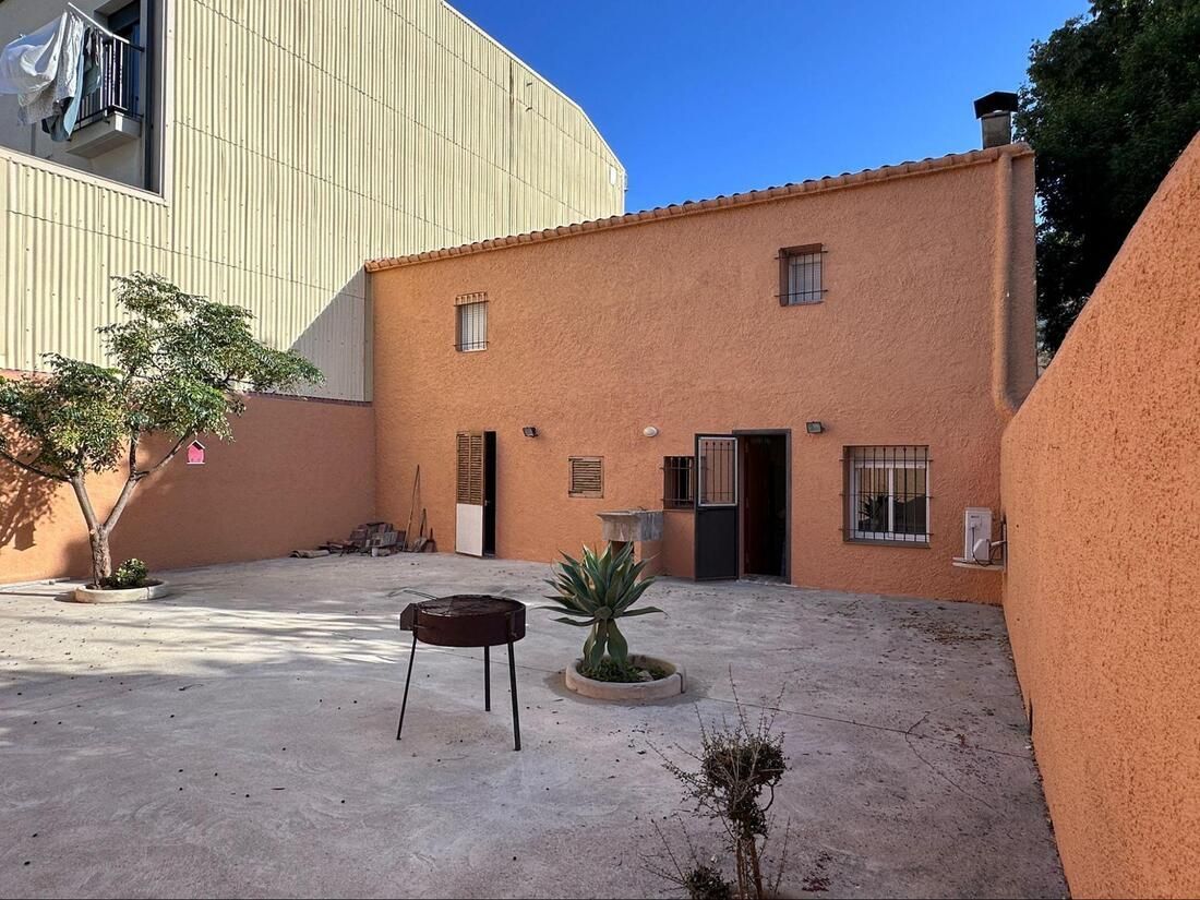 Parte trasera exterior de una casa color melocotón con un pequeño patio trasero, una barbacoa y escasas plantas.