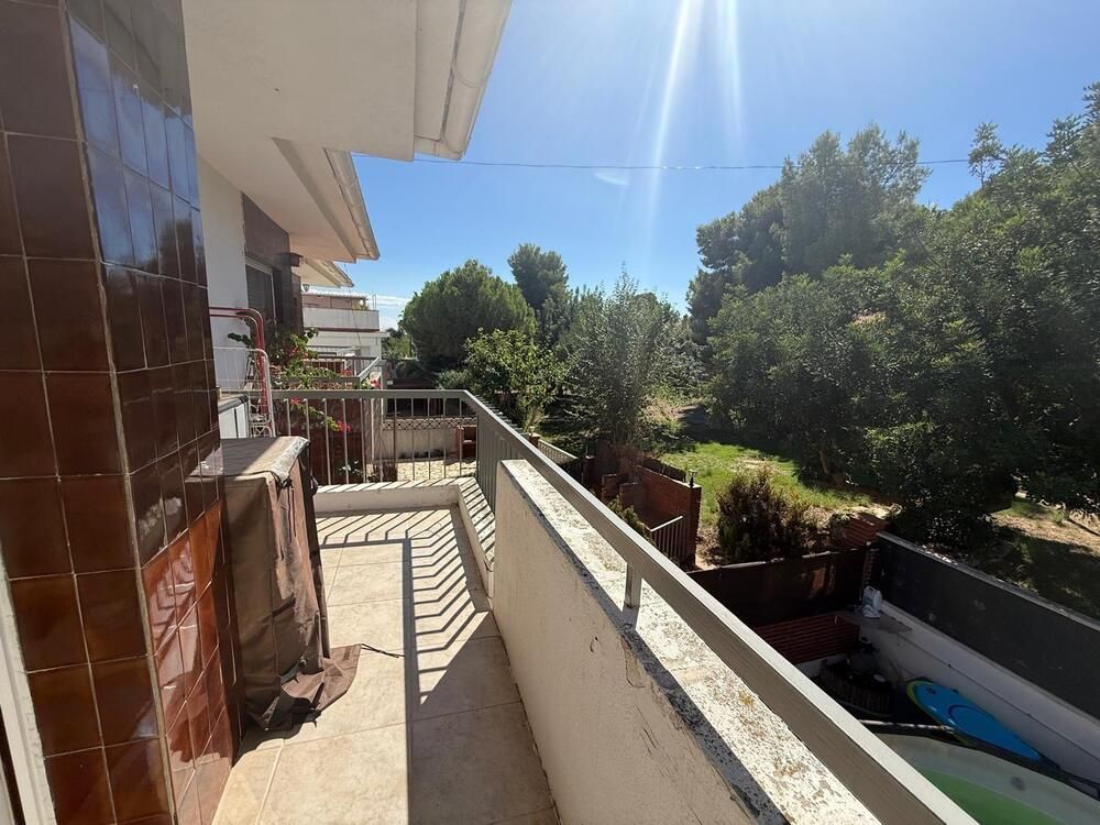 Balcón con pared de azulejos marrones, barandilla blanca, con vista a patio verde y cielo azul.
