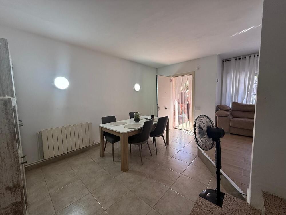 Comedor con mesa y sillas, ventilador y salida al exterior. Paredes blancas, azulejos beige y luz natural.