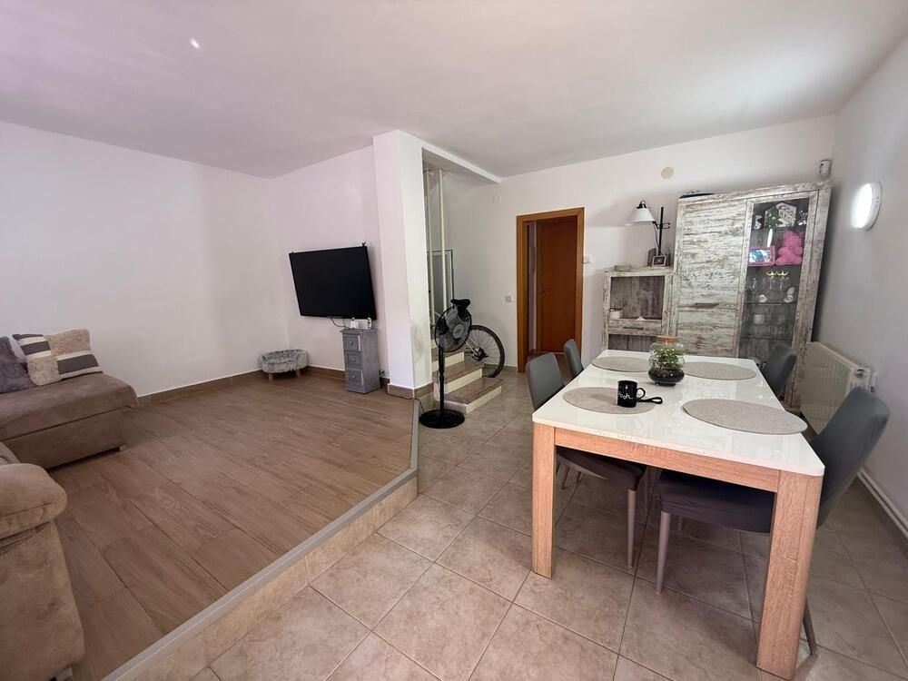 Sala de estar con comedor. Sofá marrón, televisor, mesa de madera con sillas. Escaleras con bicicleta.