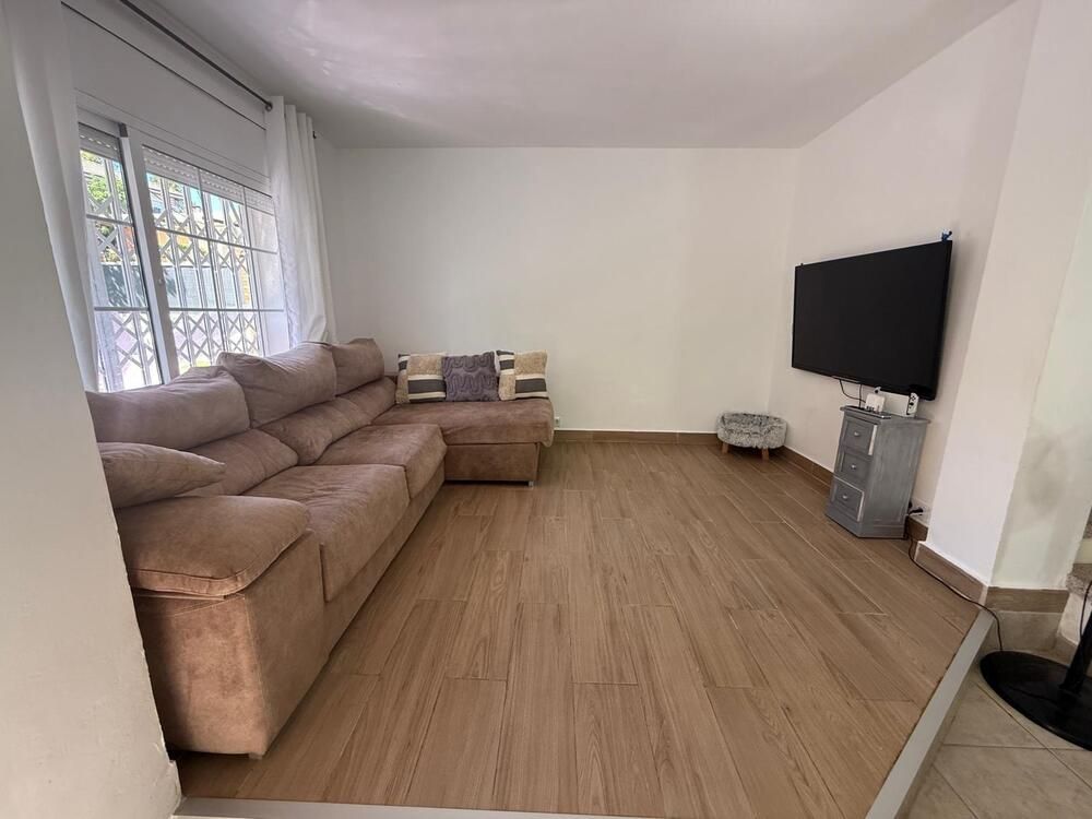 Sala de estar con sofá seccional color canela, televisor y suelo imitación madera. Paredes blancas y ventana con cortinas transparentes.