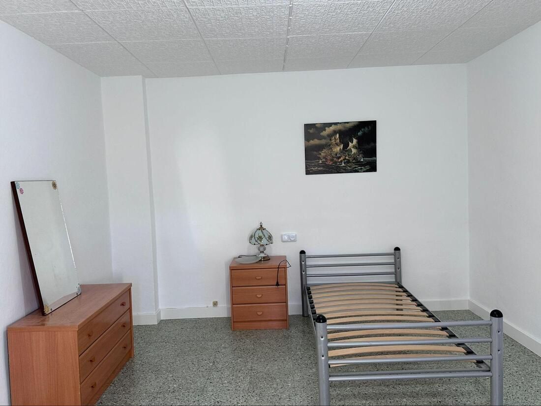 Un dormitorio escasamente amueblado con una cama, una cómoda, una mesita de noche y pequeñas obras de arte en la pared.