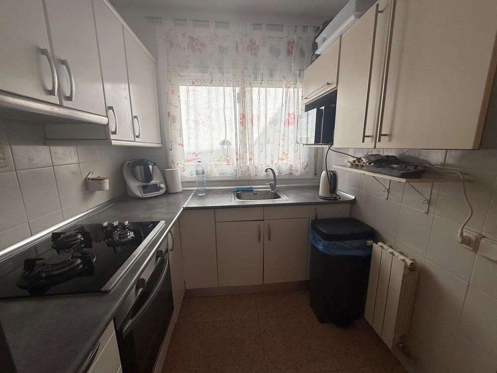 Cocina pequeña y luminosa con gabinetes blancos, electrodomésticos de acero inoxidable y una ventana con cortina transparente.