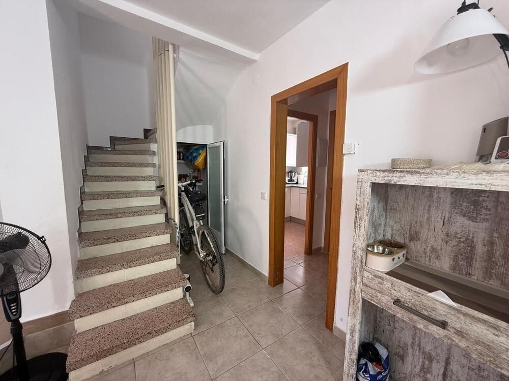 Vista interior: escalera, puerta, bicicleta y estantería. Paredes blancas, molduras de madera y suelo de baldosas.