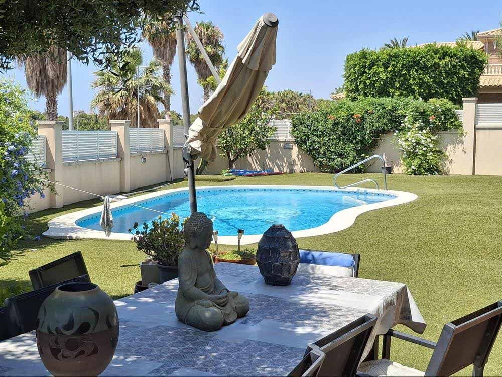 Patio trasero con piscina, mesa y estatua de Buda bajo una sombrilla en un día soleado.