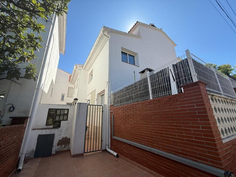 Casa adosada blanca con pared de ladrillo y portón metálico, día soleado.
