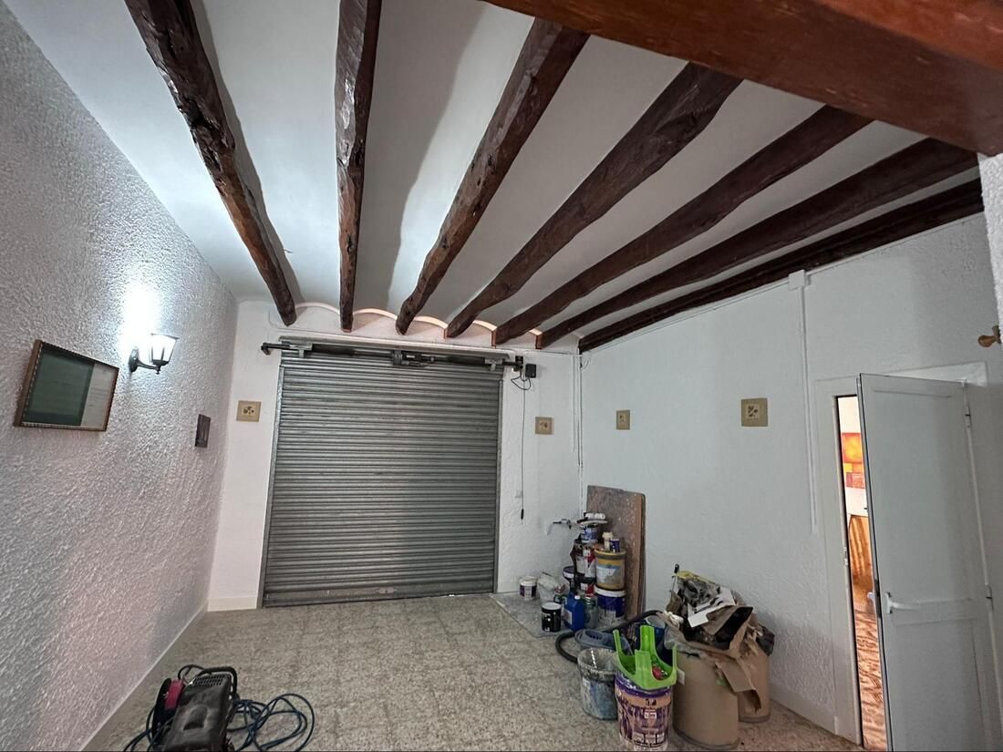 Interior de garaje con puerta enrollable, paredes blancas y vigas de madera oscura en el techo.