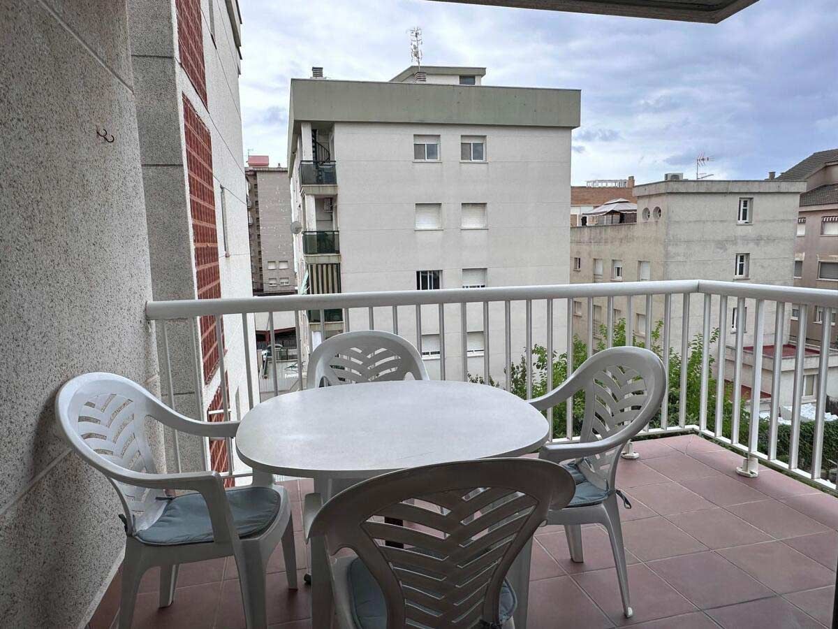Balcón con mesa blanca, sillas y vista a edificios en un entorno urbano.