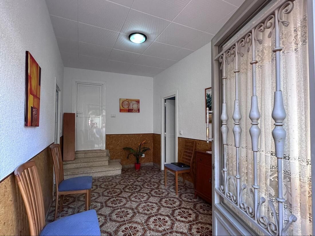 Vestíbulo con puerta metálica, suelo estampado, paredes blancas y detalles con paneles de madera; dos sillas.