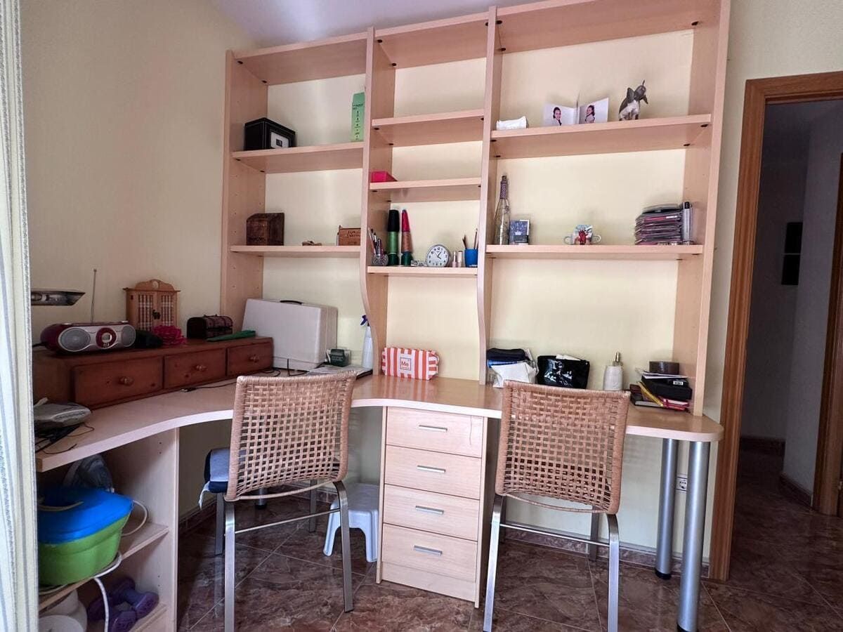 Oficina en casa con escritorio, estanterías, dos sillas y varios objetos. Paredes beige y muebles de madera.