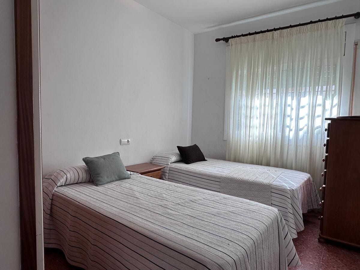 Dormitorio con dos camas individuales, paredes blancas, cortinas transparentes y una cómoda de madera.