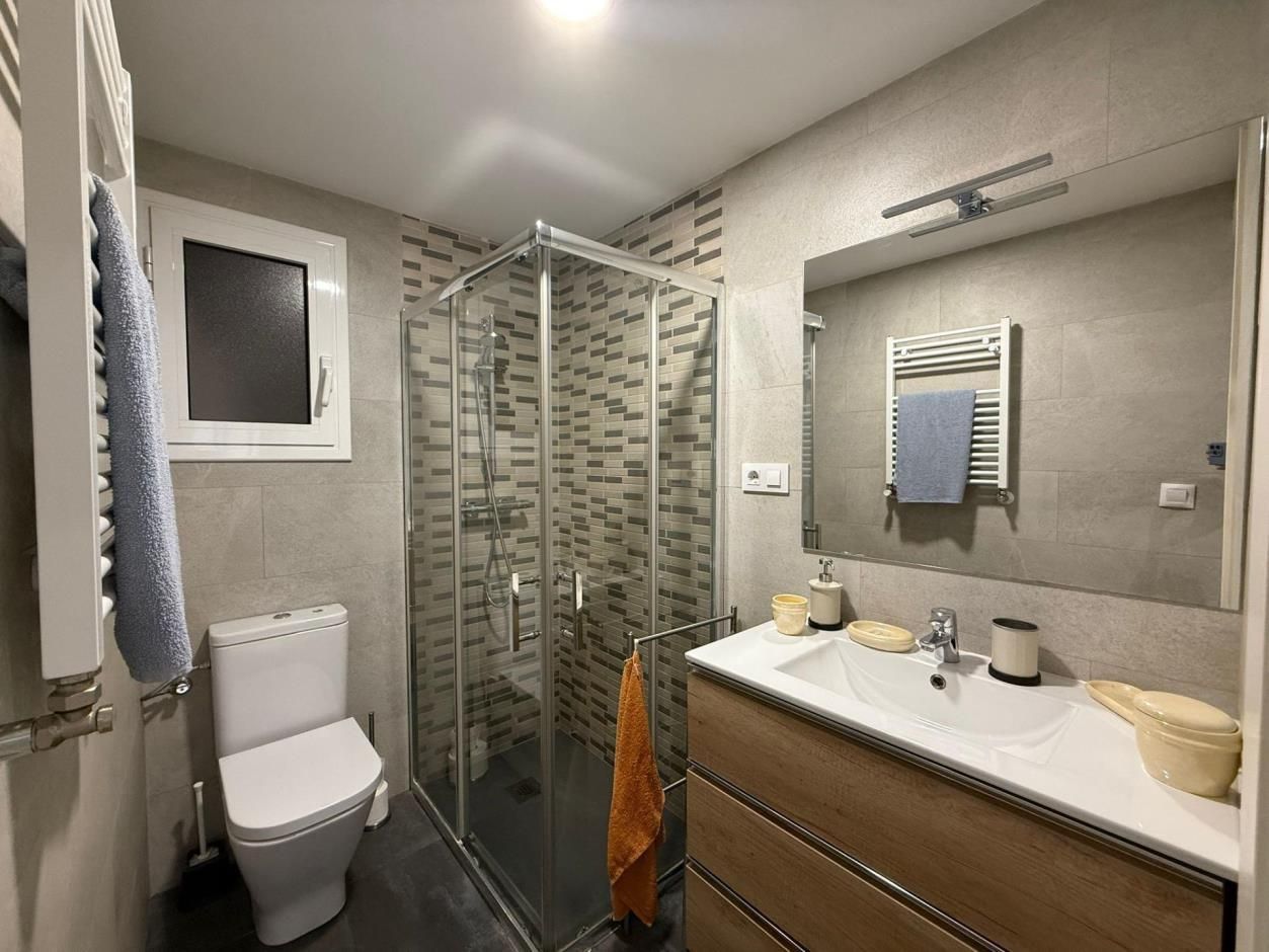 Baño moderno con ducha, inodoro, lavabo con espejo y toallero. Paredes grises.