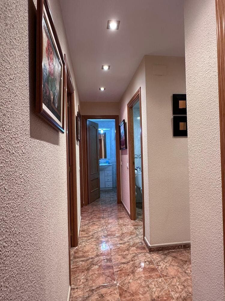 Pasillo con suelo de baldosas de color marrón rojizo, puertas a cada lado e iluminación empotrada en el techo.