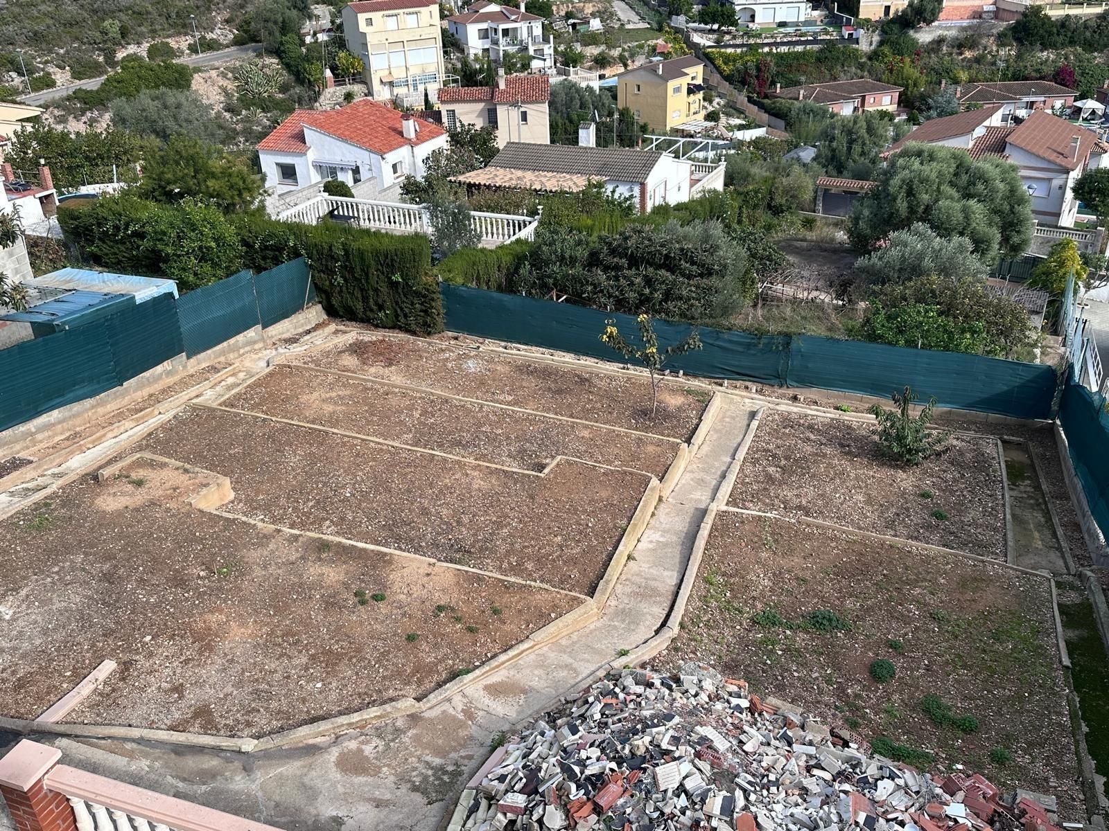 Vista aérea de un jardín de tierra dividido por senderos, rodeado por una cerca verde y edificios al fondo.