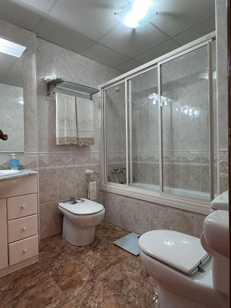 Baño con inodoro, bidé, ducha/bañera combinada, tocador y toallero. Combinación de colores beige y marrón.