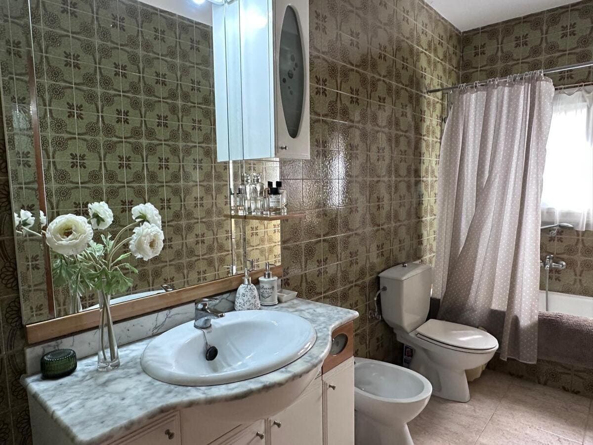 Baño con azulejos verdes con estampado floral, lavabo blanco, inodoro, bidé y cortina de ducha.
