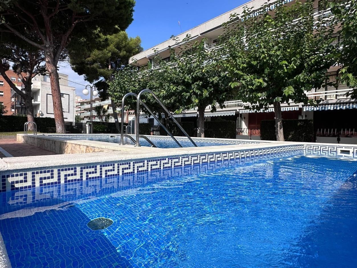 Piscina con agua azul y borde de azulejos azules y blancos; árboles y edificios al fondo.