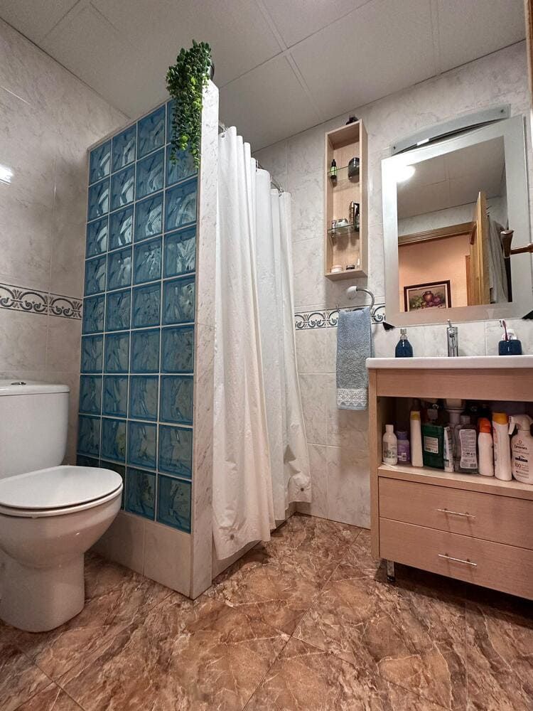 Baño con ducha de bloques de vidrio, inodoro, lavabo, mueble y azulejos de color marrón/bronceado.