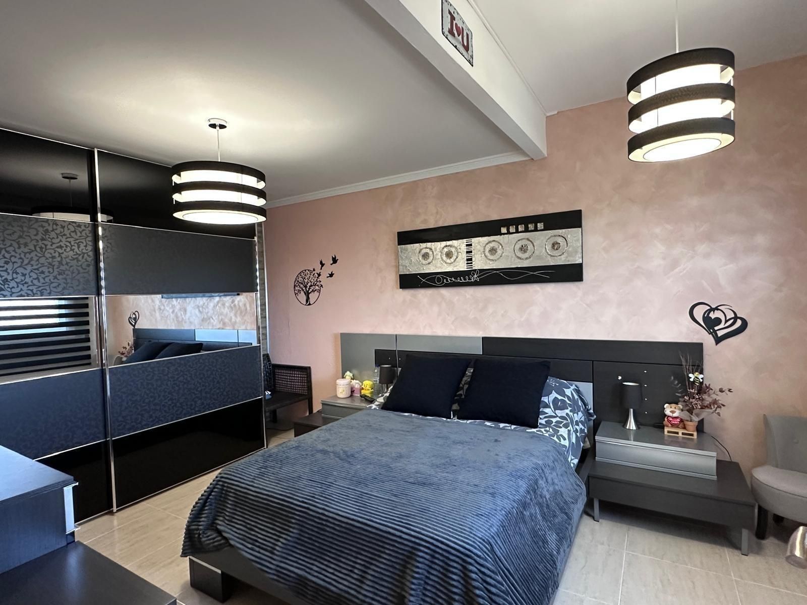 Dormitorio con cama, mesitas de noche, armario y lámparas colgantes. Paredes rosas, muebles oscuros y una obra de arte con una cinta de casete.