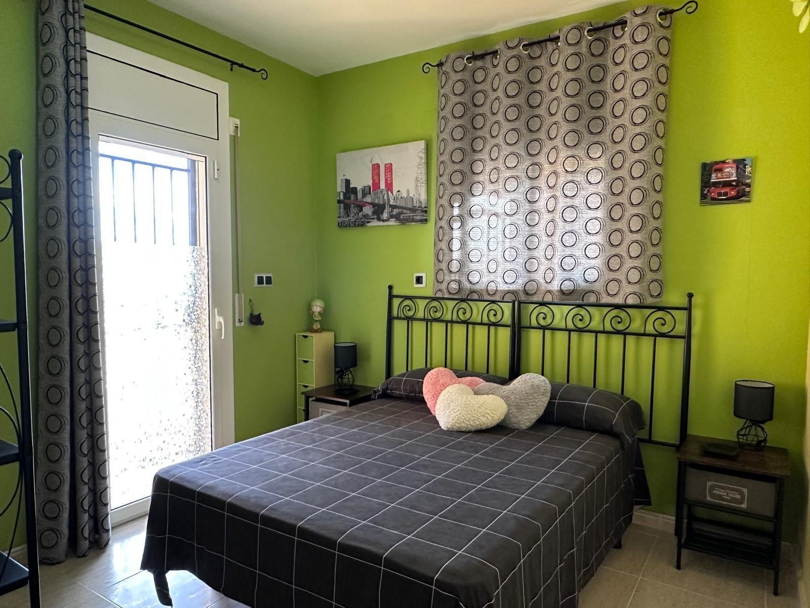 Dormitorio con paredes verdes, marco de cama de metal negro, ropa de cama con estampado oscuro y puerta de balcón.