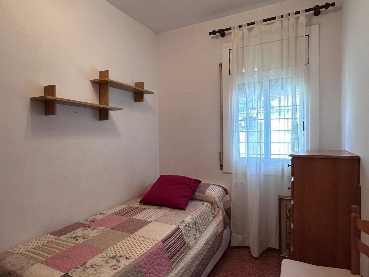 Dormitorio con una cama individual, pequeña cómoda, estanterías y una ventana con cortinas transparentes.