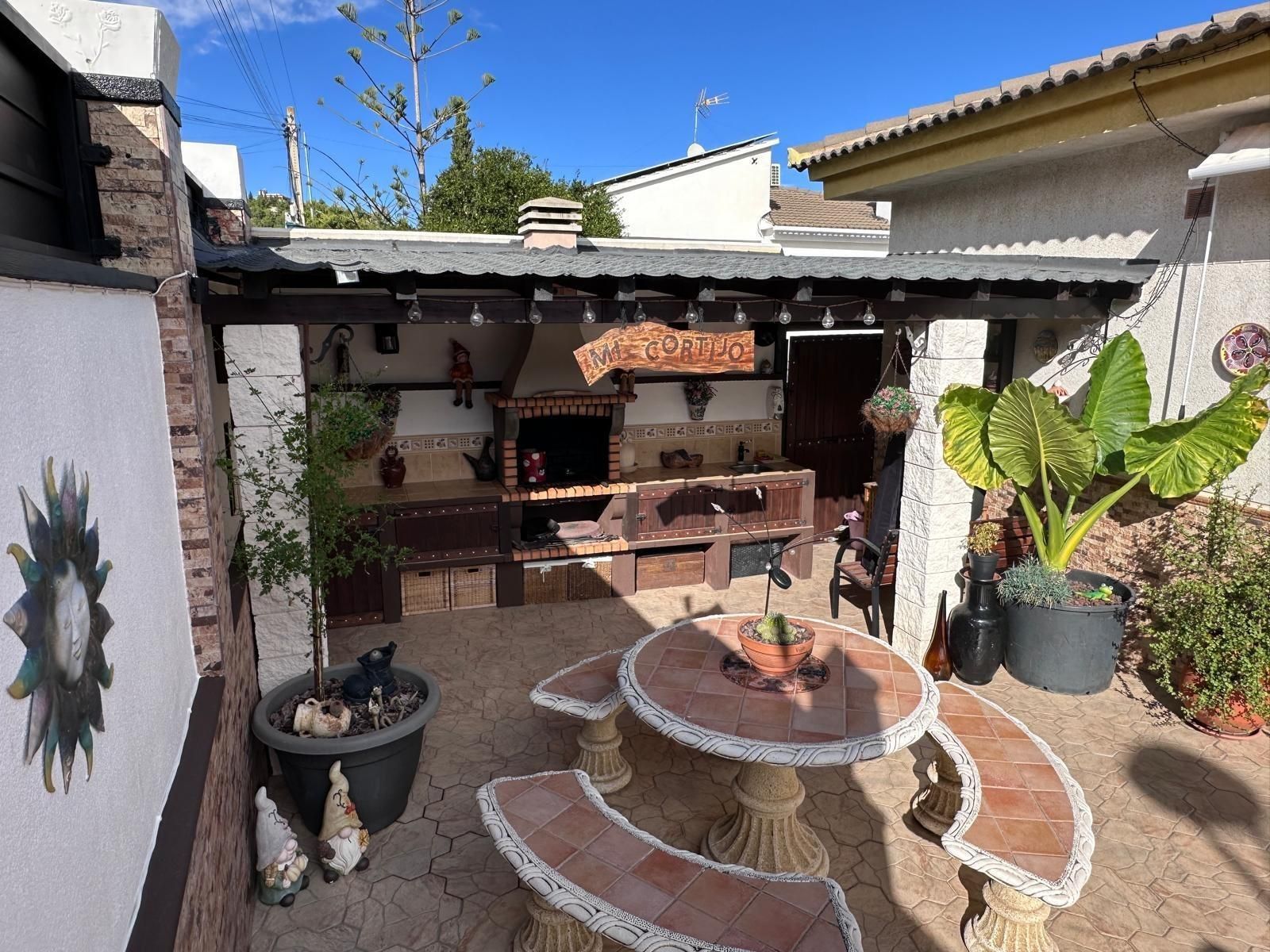 Patio exterior con barbacoa incorporada, mesa y plantas en macetas bajo techo de chapa ondulada.