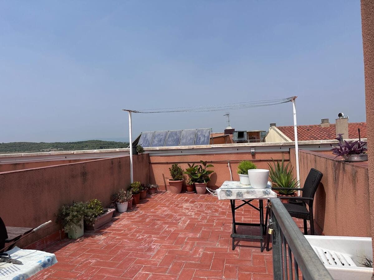 Terraza en la azotea con ladrillos rojos, plantas, muebles y una cadena montañosa lejana bajo un cielo azul.