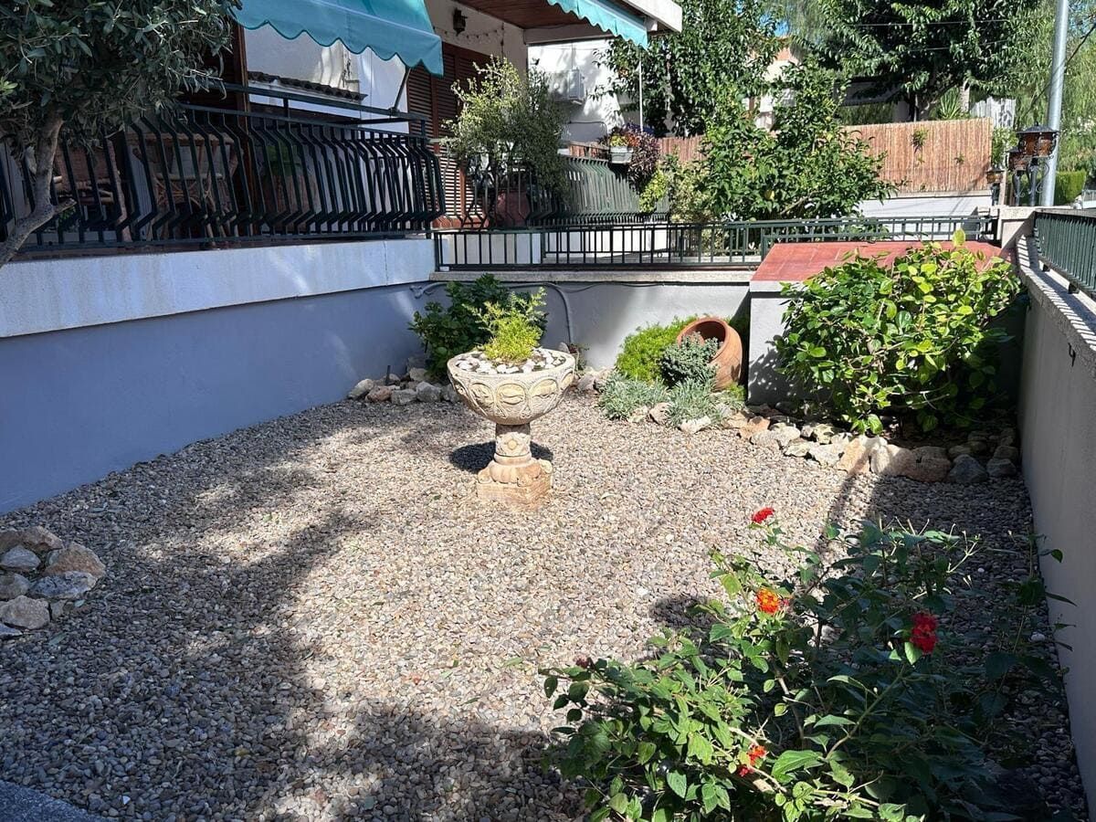 Pequeño jardín con grava, urna de piedra y arbustos delante de un edificio.