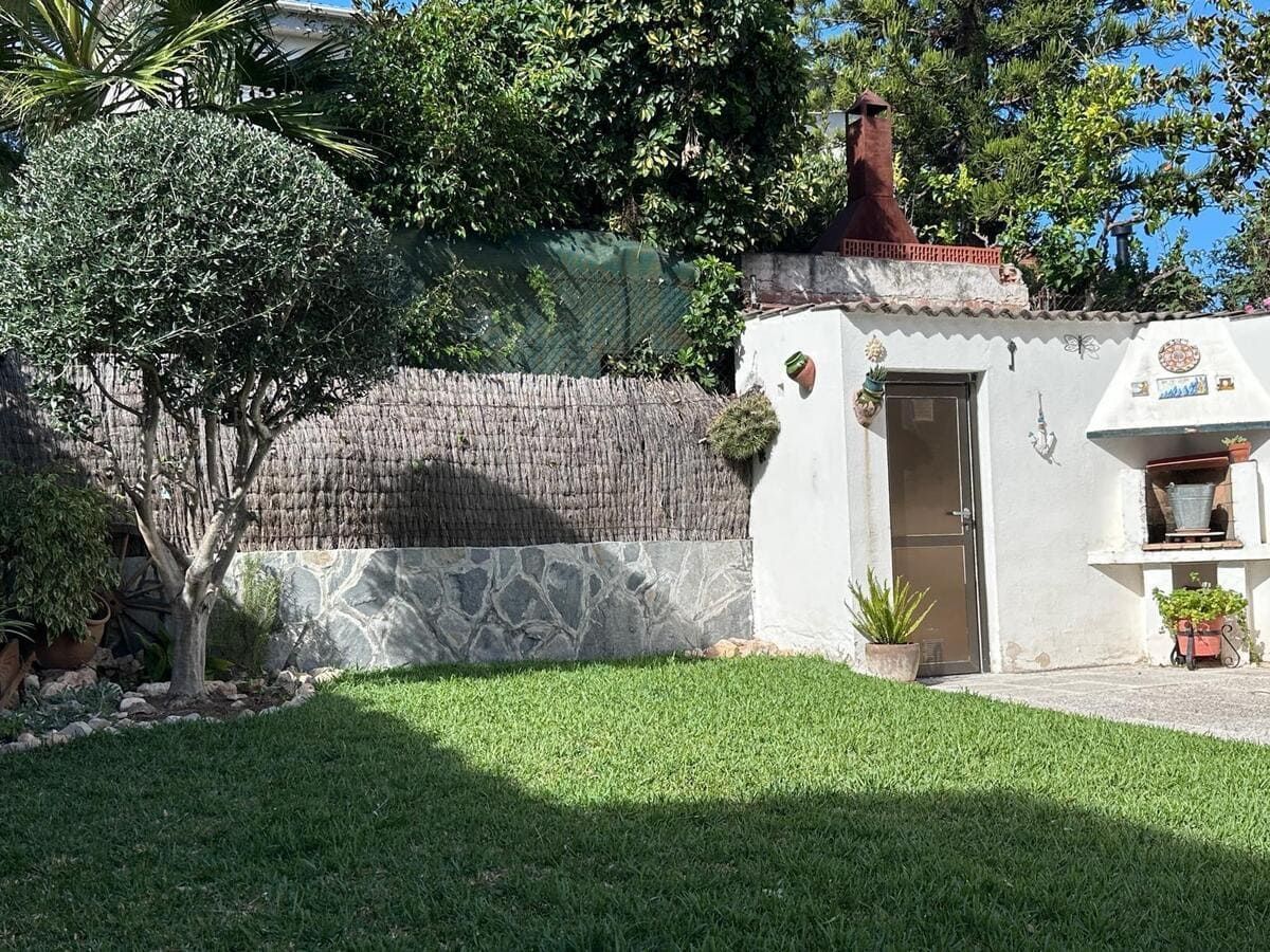 Césped verde con un árbol, un muro de piedra decorativo y un edificio de estuco blanco con puerta y chimenea.