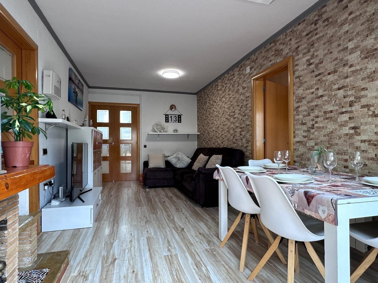 Sala de estar con sofá oscuro, mesa de comedor para invitados, pared con acento de ladrillo y piso de madera clara.