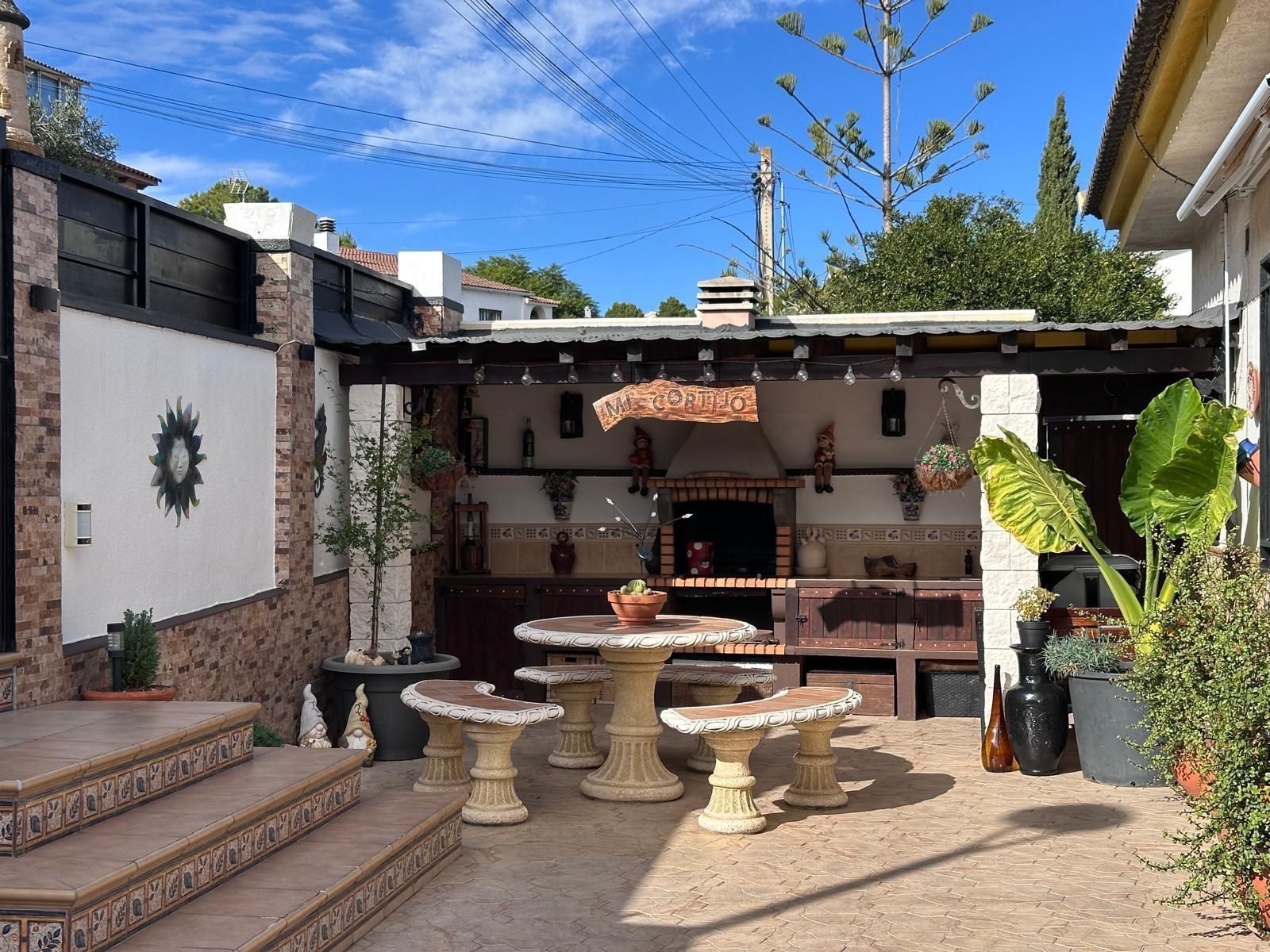 Patio exterior con mesa de piedra, bancos y barbacoa de obra bajo marquesina.