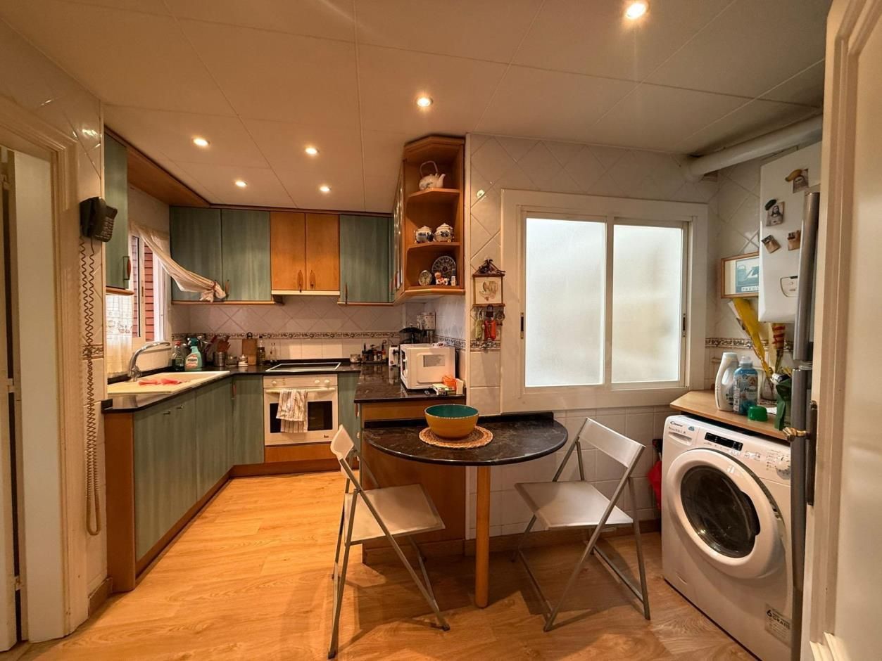 Cocina pequeña con armarios color verde azulado, suelo de madera y mesa redonda con dos sillas. Incluye ventana y lavadora.