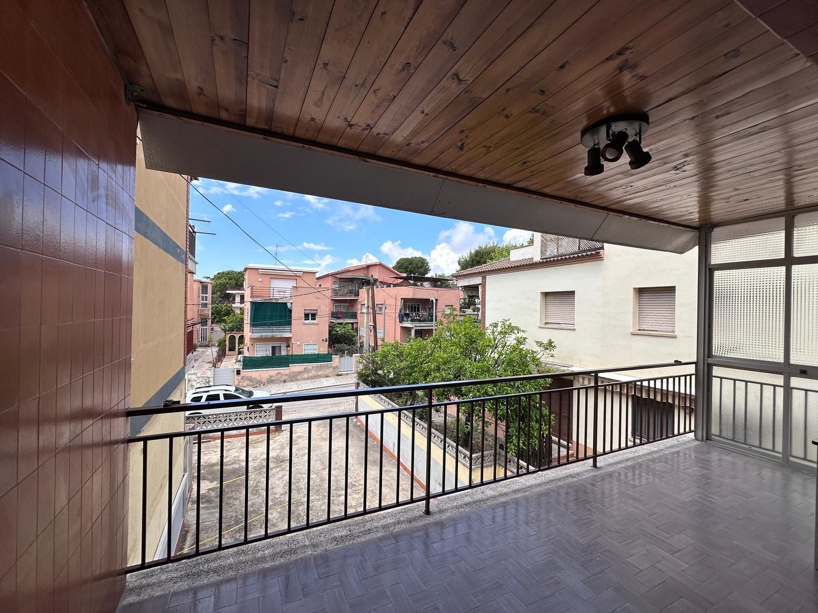 Balcón con vista a edificios; barandilla negra, techo de madera, cielo despejado.