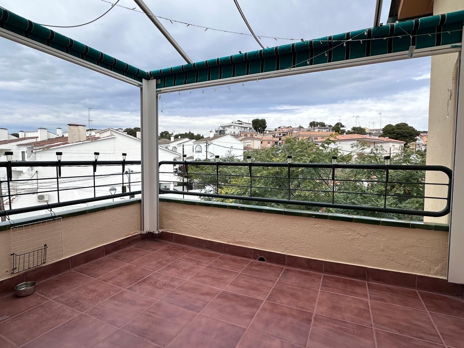 Balcón con piso de baldosas rojas, barandilla negra, toldo y vista a edificios bajo cielo nublado.