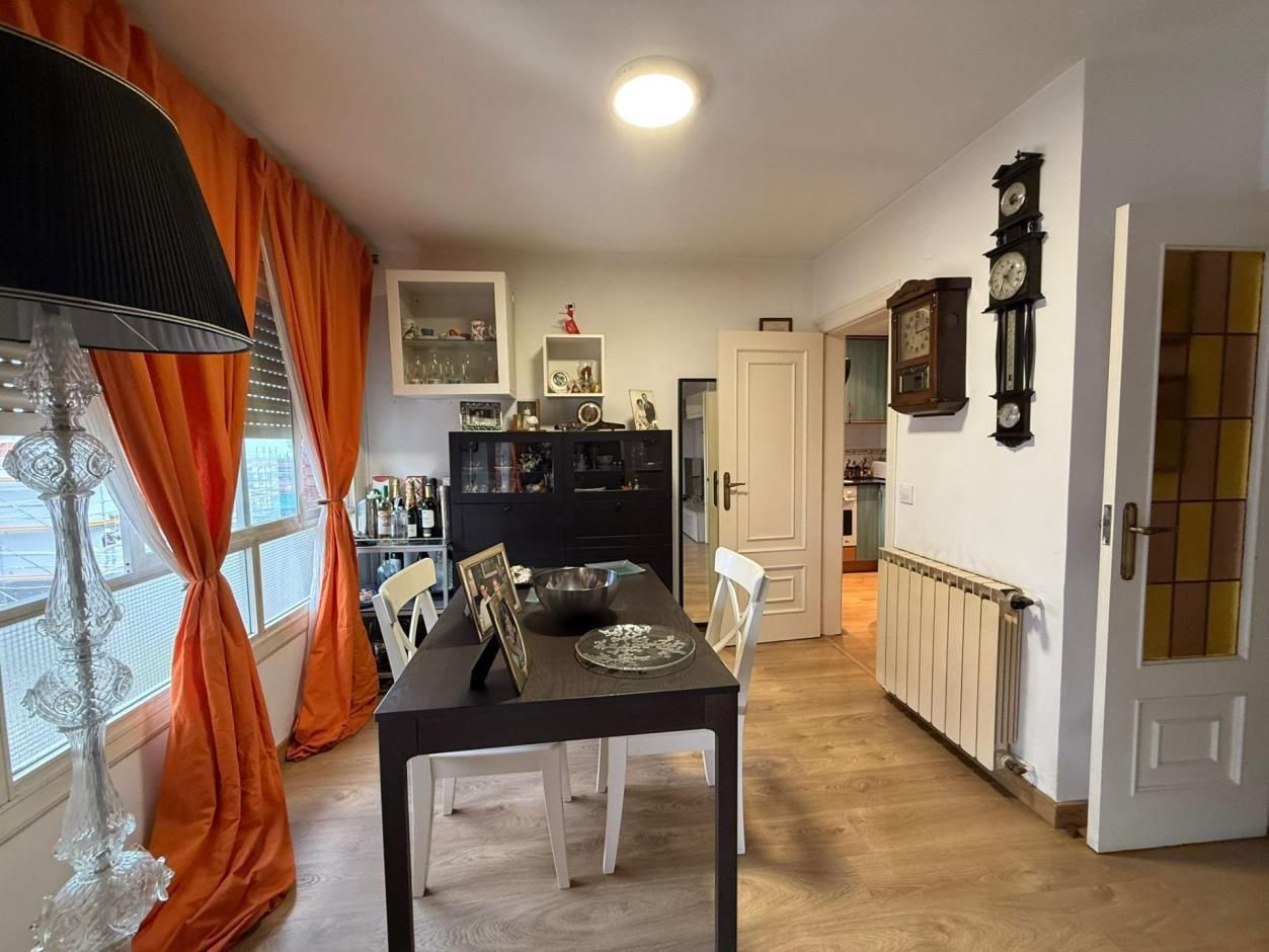 Comedor con cortinas naranjas, mesa oscura y sillas blancas. Suelo de madera, armarios y reloj.