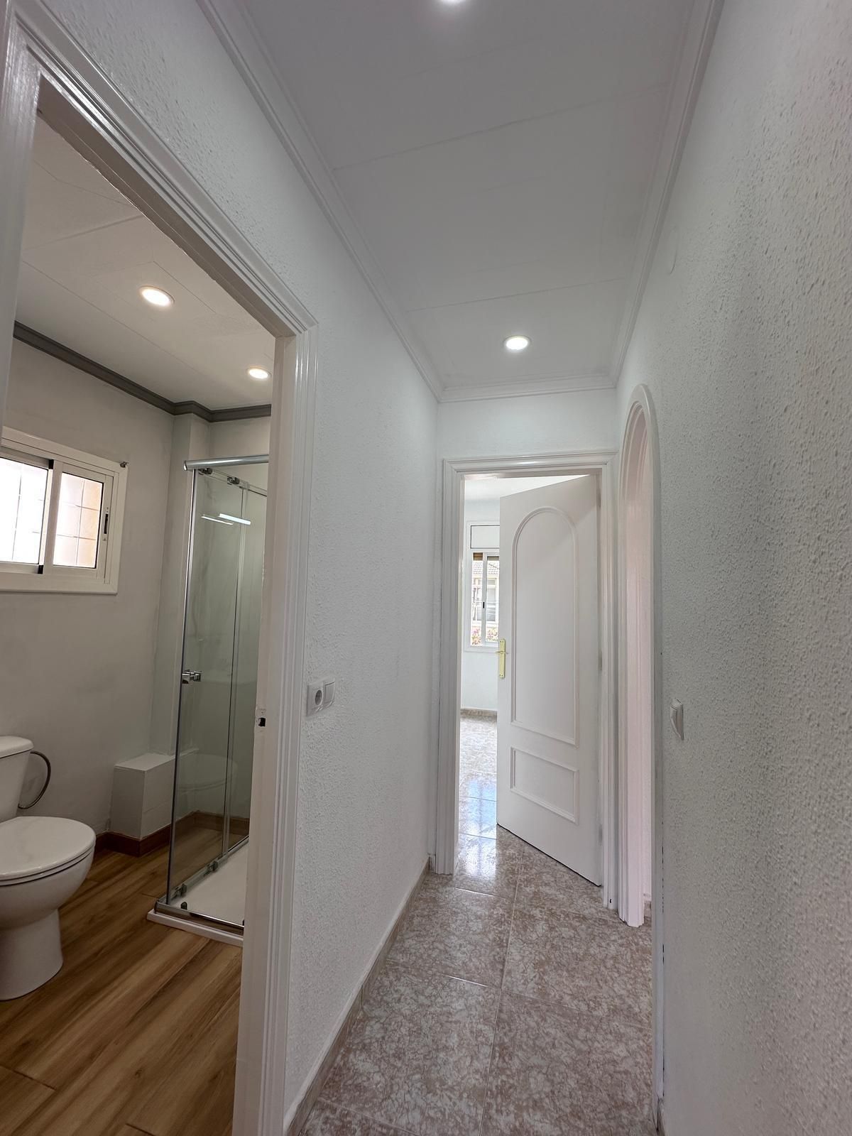 Un pasillo luminoso con un baño a la izquierda y una habitación al fondo. Paredes blancas, suelo de madera y puertas abiertas.