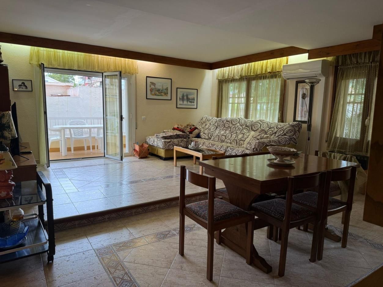 Sala de estar con mesa de comedor, sofá y acceso al balcón. Luz natural, tonos cálidos y telas estampadas.