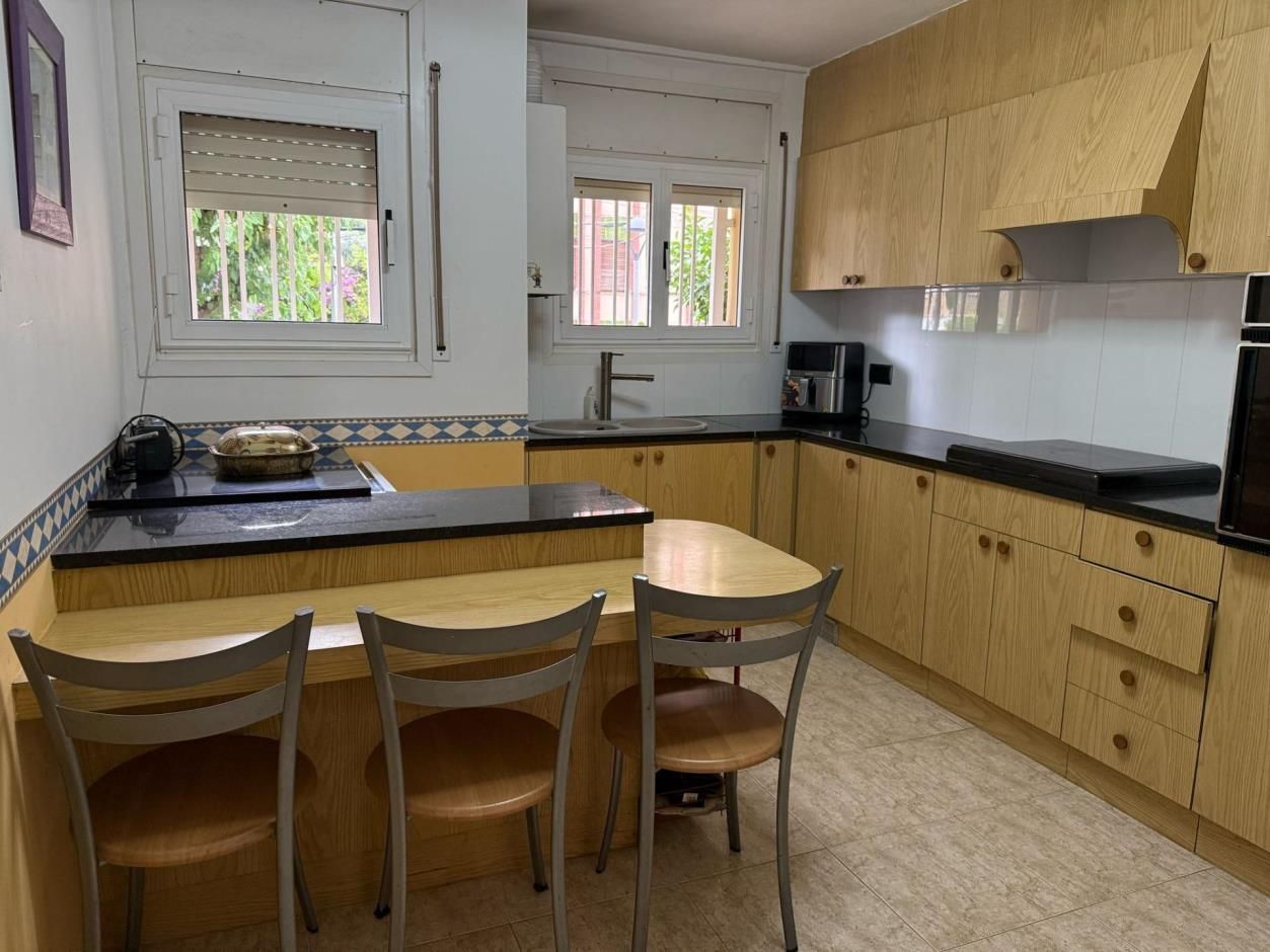 Cocina con gabinetes de madera clara, barra desayunadora con sillas, ventanas y encimeras negras.